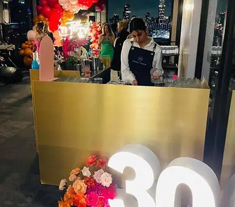 Bartender behind a gold bar beside a lit-up 30 sign at a 30th birthday party
