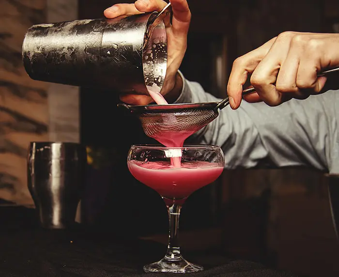 Bartender double-straining a pink cocktail into a coupe