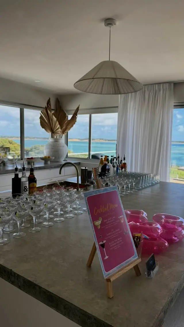 Cocktail glasses and a pink cocktail menu on a kitchen bench with a sea view